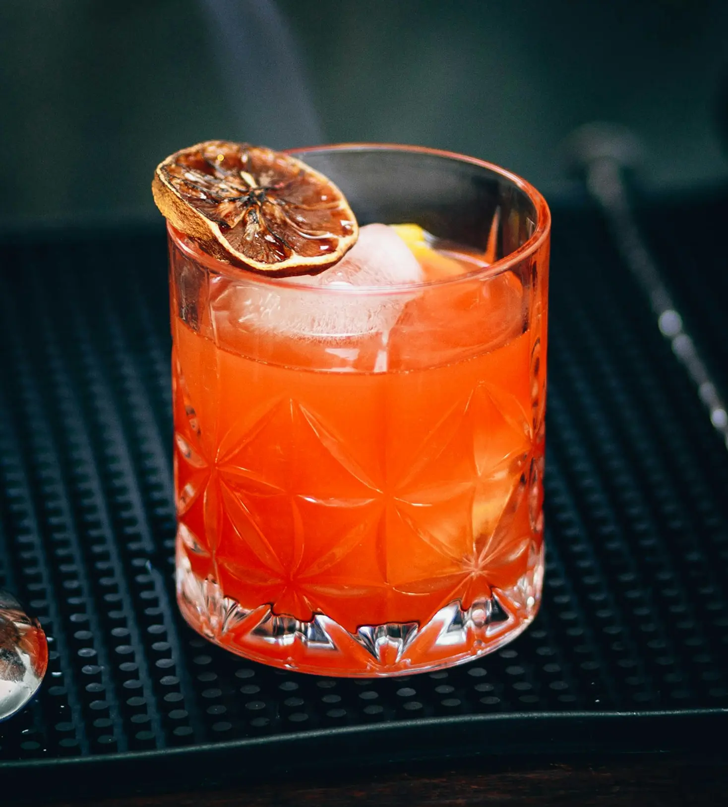 Cocktail arancione con fetta di agrume essiccato su un bancone scuro.