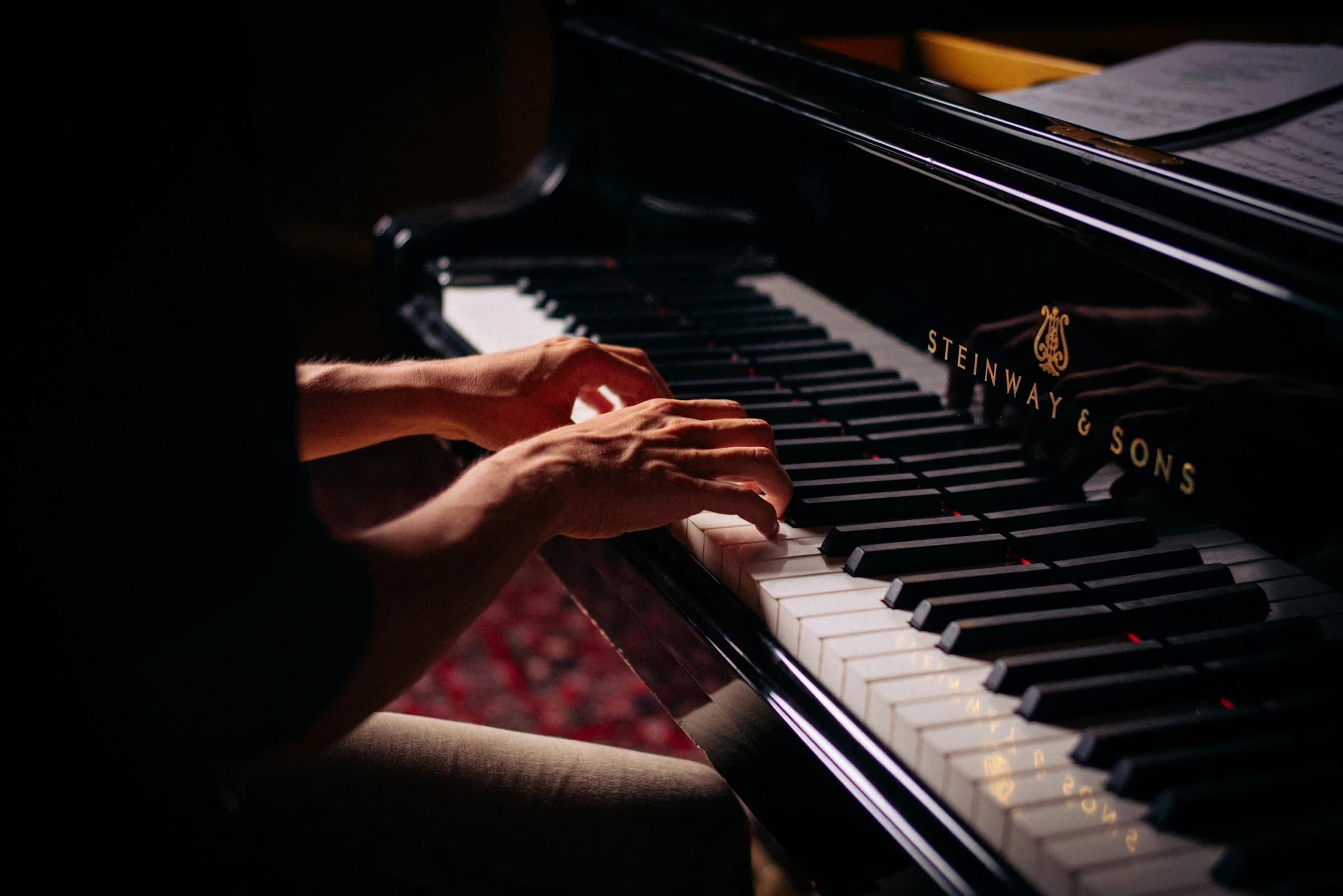 Primo piano di mani che suonano un pianoforte a coda in un locale in penombra.