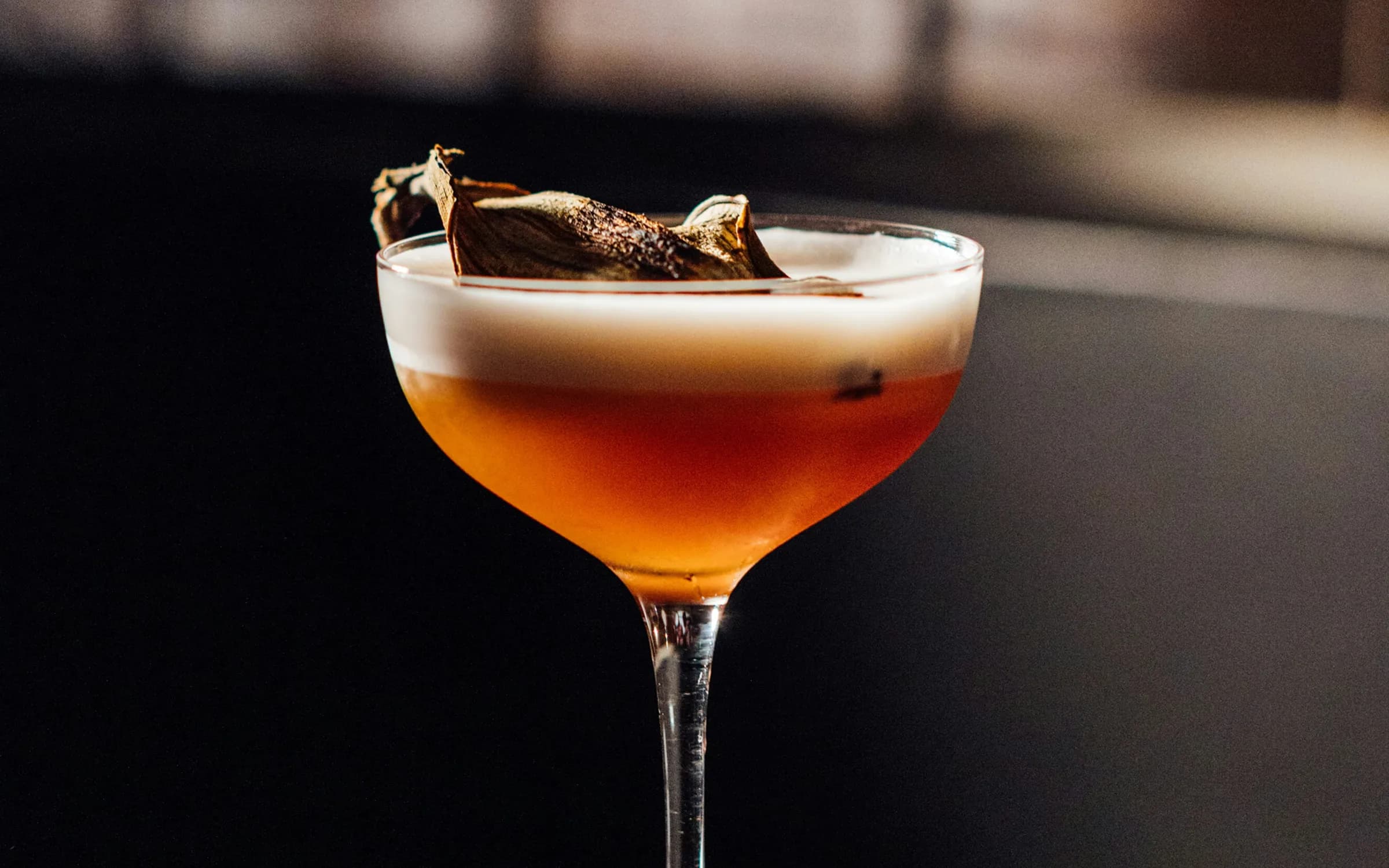 Cocktail ambrato in coppa con schiuma bianca e una foglia essiccata come guarnizione, su sfondo scuro.