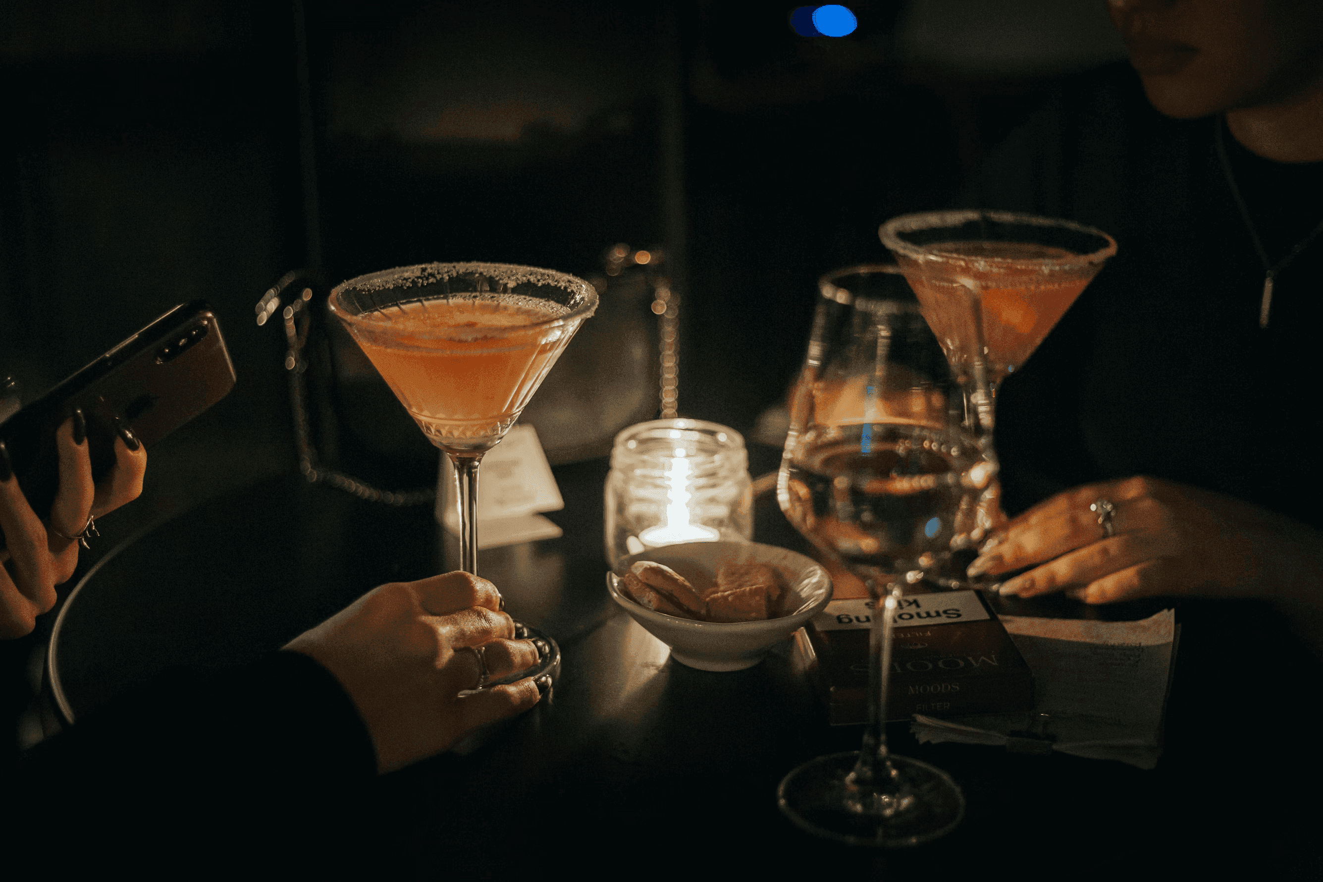 Due cocktail rosa con bordo salato su un tavolo, tra candele e un pacchetto di sigarette.